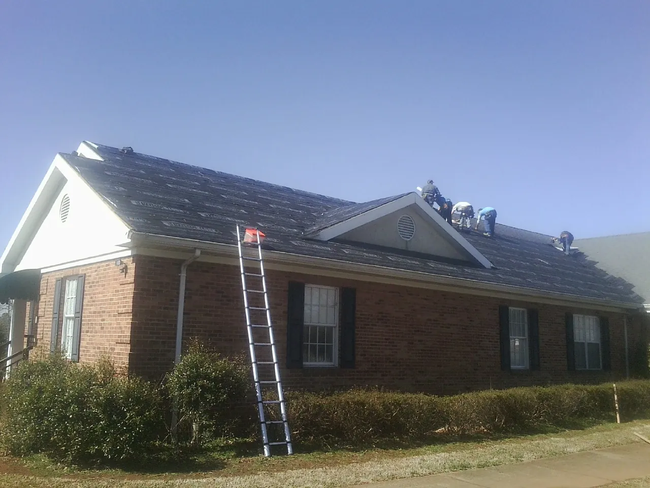 Crew tearing shingles off a brick ranch roof with a ladder set — Emergency Roof Repair work by our crew in Pine Lake
