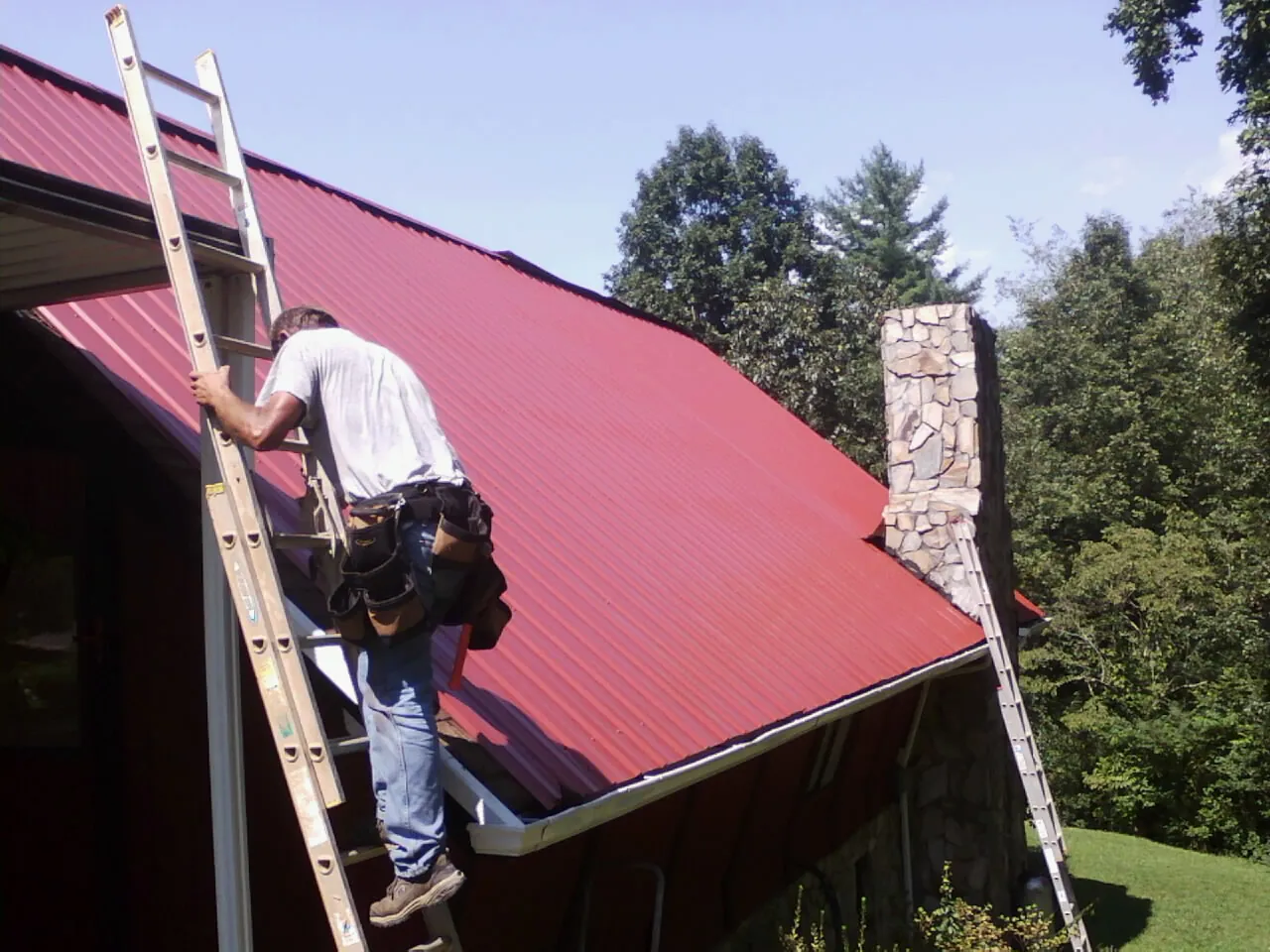 Roofer on a ladder beside a red metal roof and stone chimney — Soffit & Fascia Repair by Roofing Brothers in Pine Lake